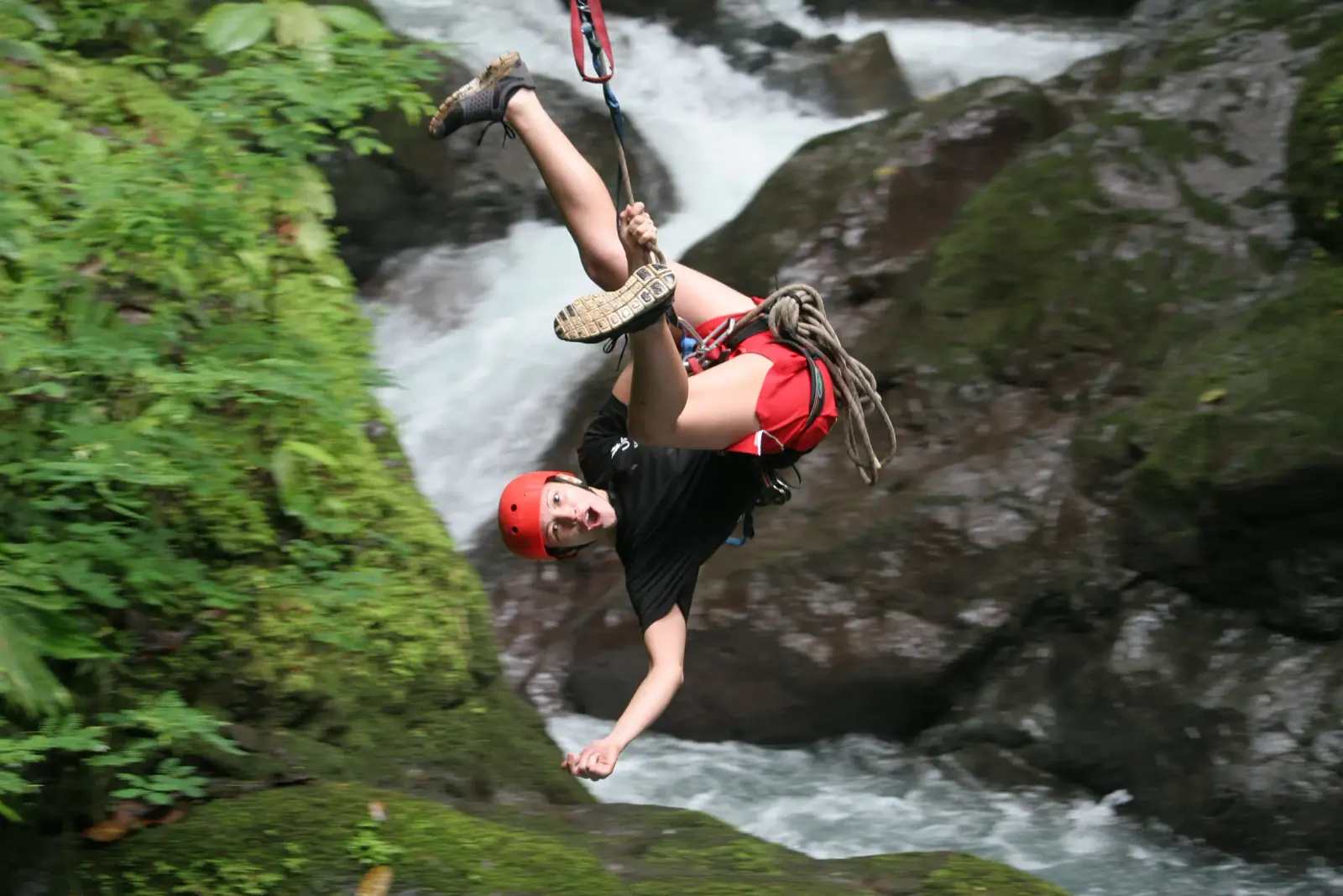 Rappeller in a red helmet hanging upside down beside a jungle waterfall