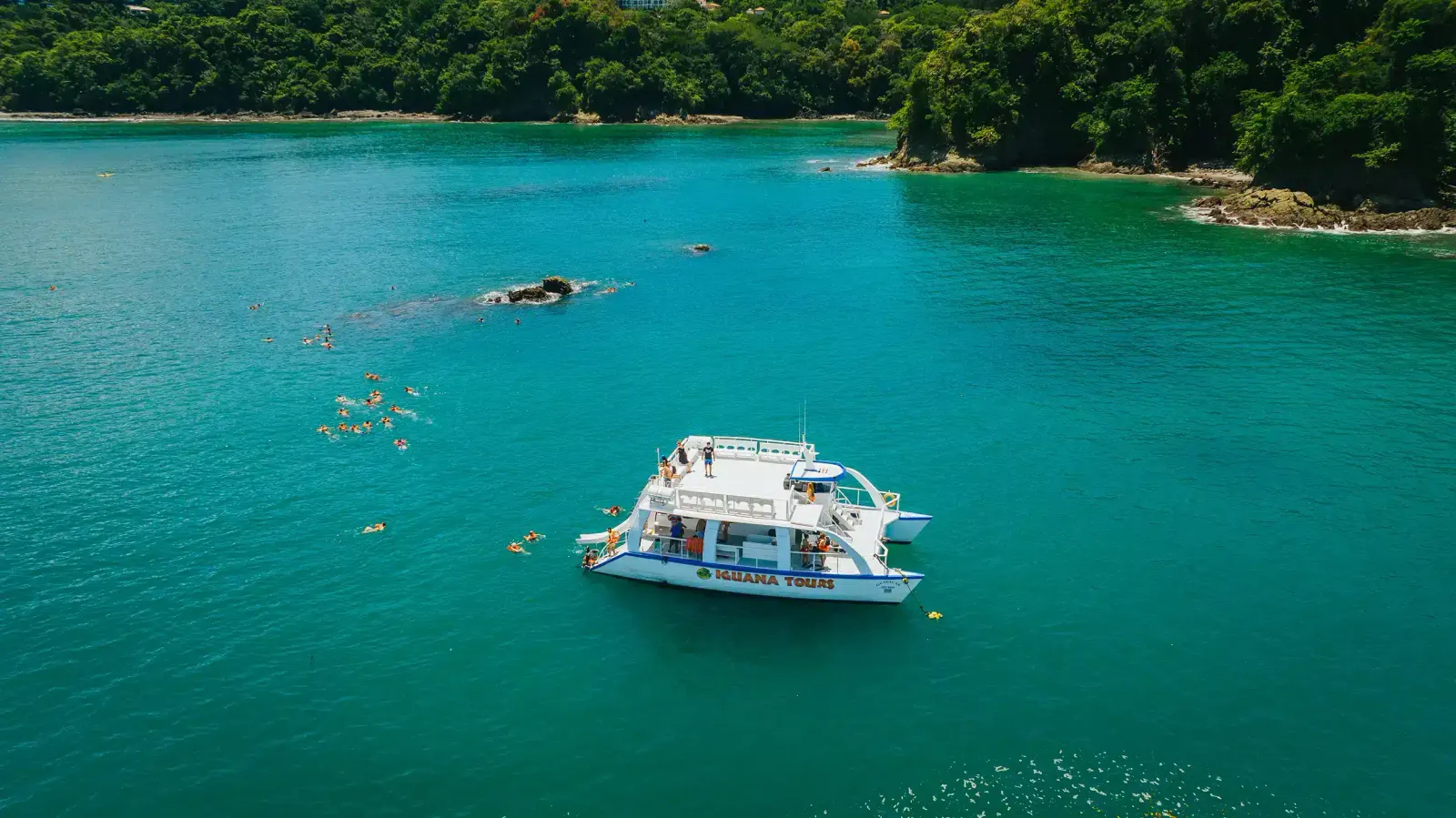 Aerial view of a tour catamaran anchored in a turquoise bay with snorkelers in the water