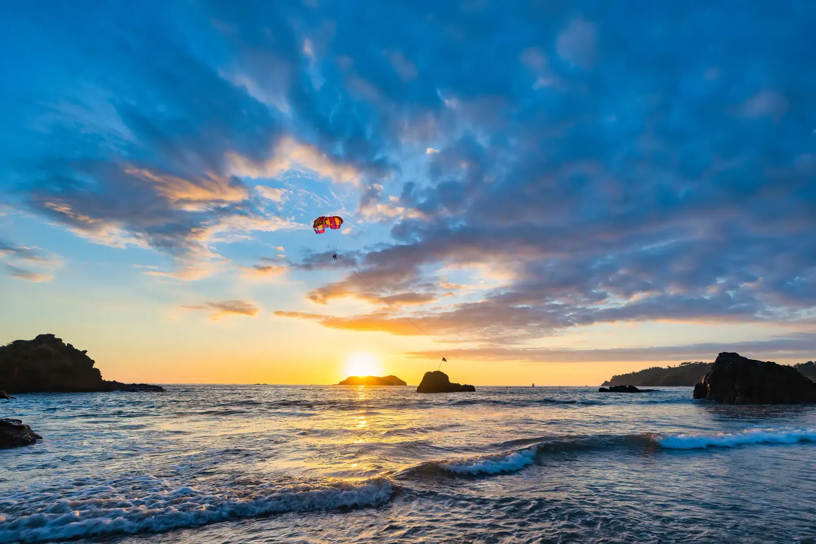 Red parasail high over the Pacific at sunset, with rocky islets on the horizon