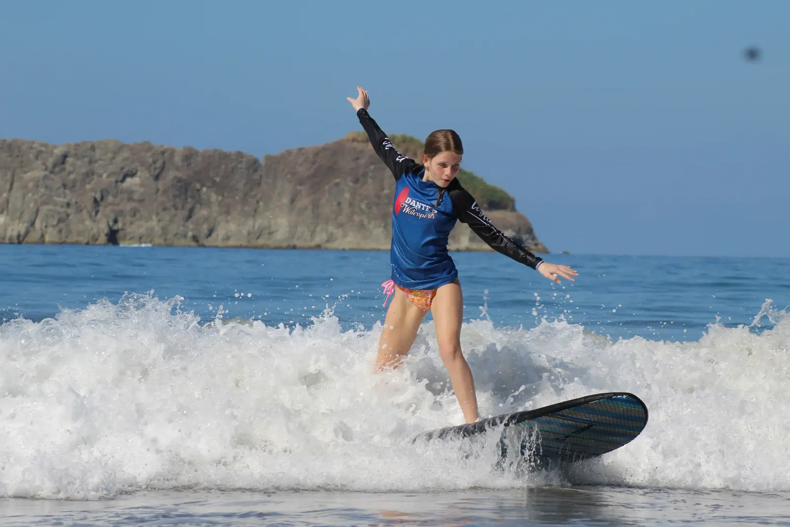 Girl in a rash guard riding a surfboard on a small breaking wave, with a rocky headland behind