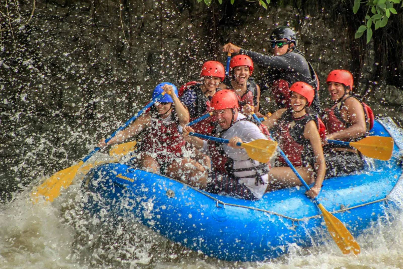 Rafters in red helmets paddling a blue raft through splashing whitewater