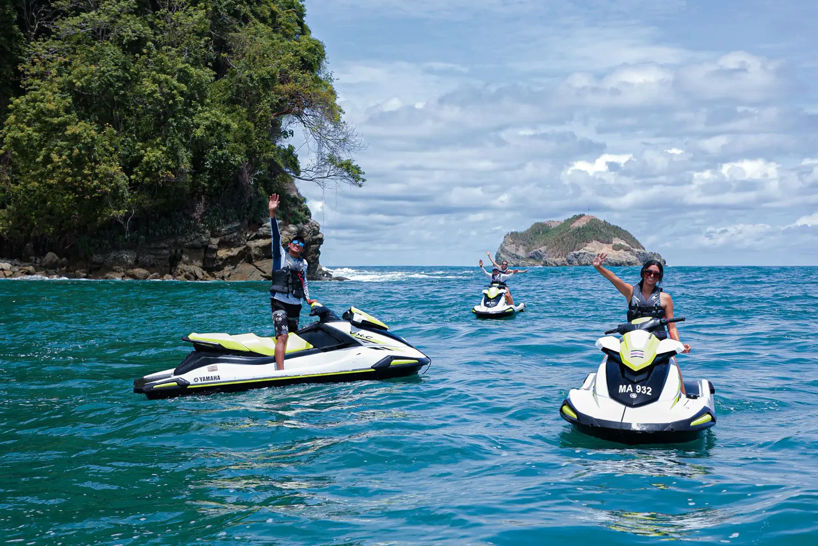Two riders standing on jet skis with arms raised, near a rocky islet