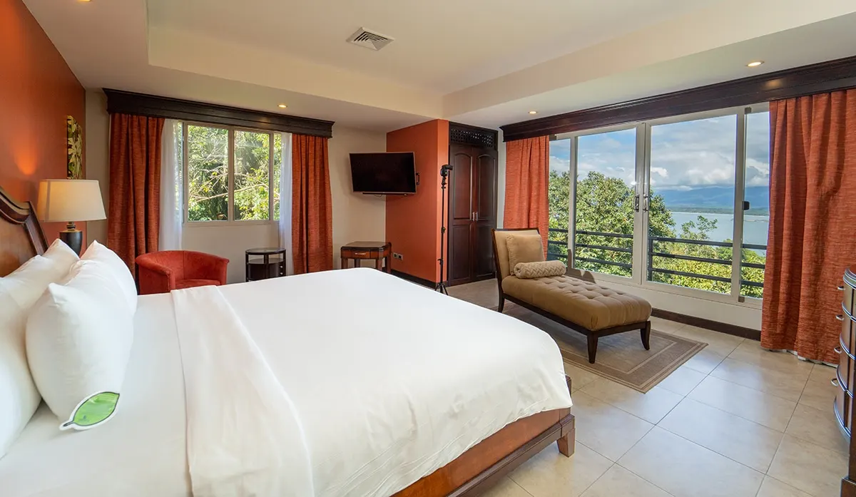 Penthouse bedroom with a king bed in white linens, a chaise longue by floor-to-ceiling windows, and the forest and ocean beyond the balcony rail