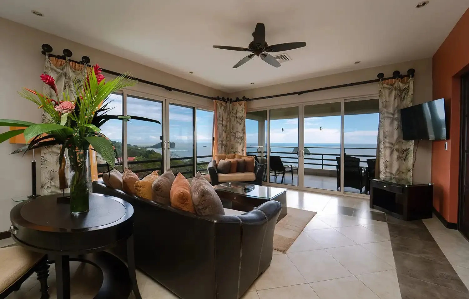 Master Vista Suite living room with a leather sofa, a vase of red ginger and heliconia, and a wall of sliding glass doors open to the Pacific and the Manuel Antonio coast