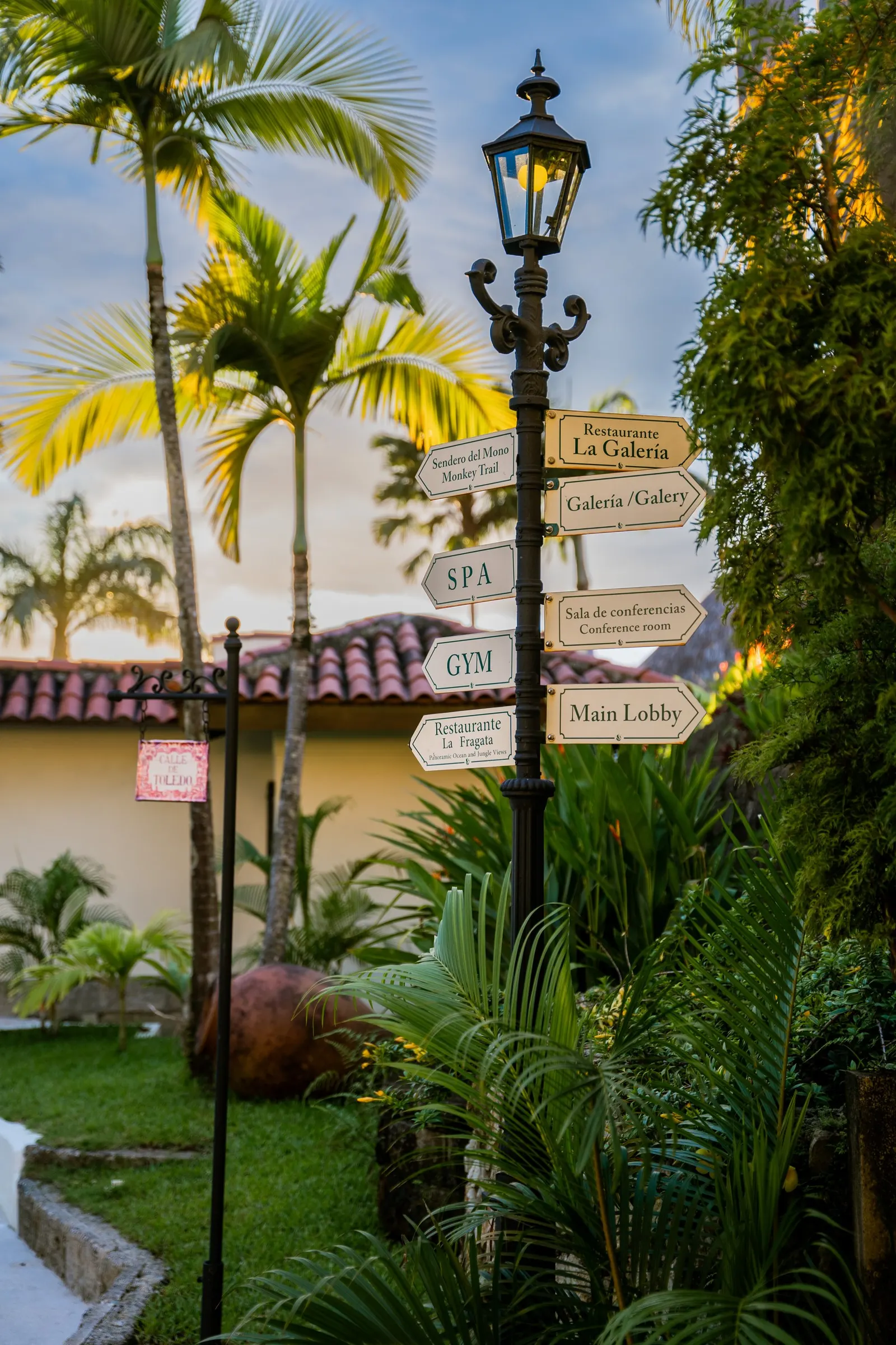 An iron lamppost at sunset with signs to the Monkey Trail, La Galería, the spa, the gym, La Fragata and the main lobby, palms and a tiled roof behind